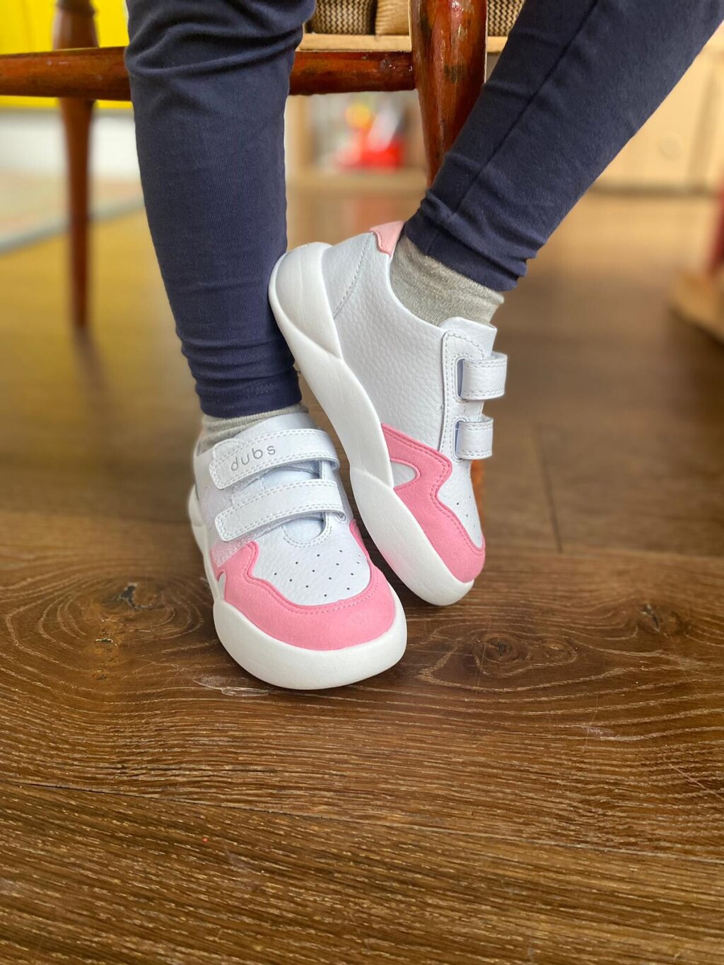 A close-up image of a pair of children's white and pink trainers feature central on a wooden floor with wooden chair legs to the back. Dubs branding is written on the white leather strap.