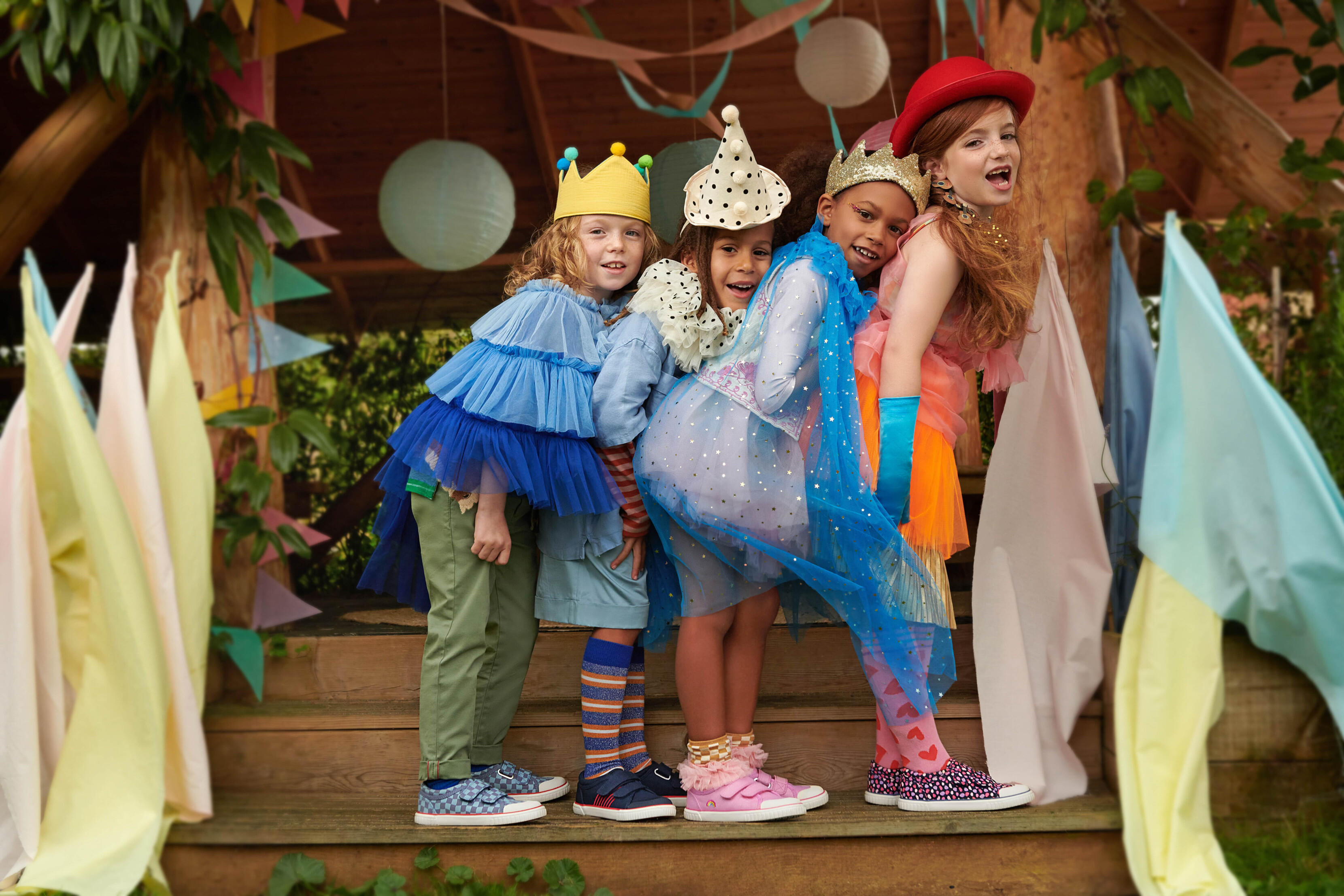 Marketing image by Start Rite Shoes. Four children feature central on wooden tree house steps. From left, a child wears a yellow crown, blue tutu, and blue plimpsoles, second a pair of blue and red trainers with striped socks and a white and black spotted party hat, third from left features a girl wearing a gold crown, blue tulle cape, and a pair of pink trainers, to right a girl with auburn hair wears a red bowler hat, orange kirt and bright spotted shoes. All children and posed smiling at the camera and flags feature in the foreground.