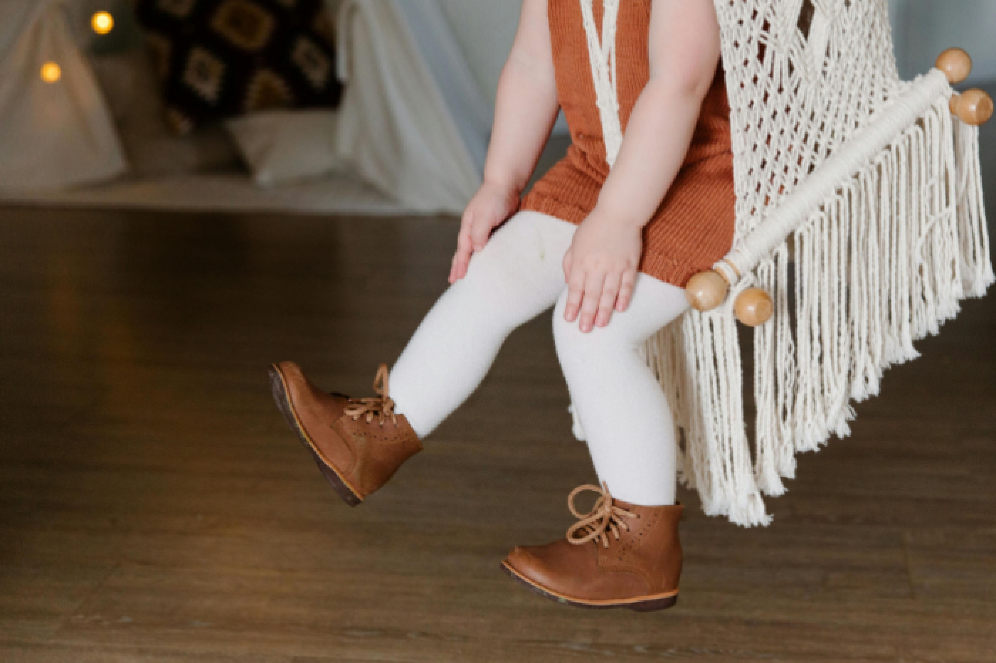 Image shows a child dressed in a tan cord dress, white woolen tights and brown leather laced boots, swinging on a macrame seat against a wooden floor and white clothed background.