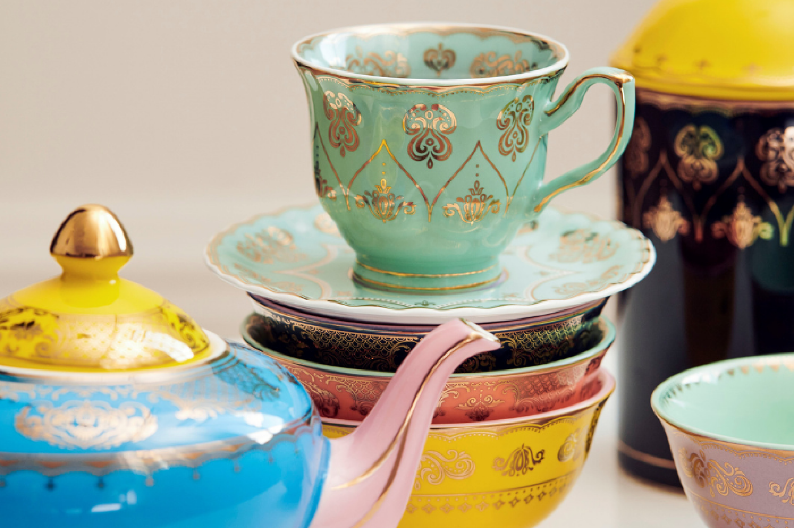 Extreme close up of bright coloured tableware with a mint green and gold teacup and saucer, a yellow, blue and pink teapot, and black and yellow vase to the top right of image. Image supplied by brand POLSPOTTEN.