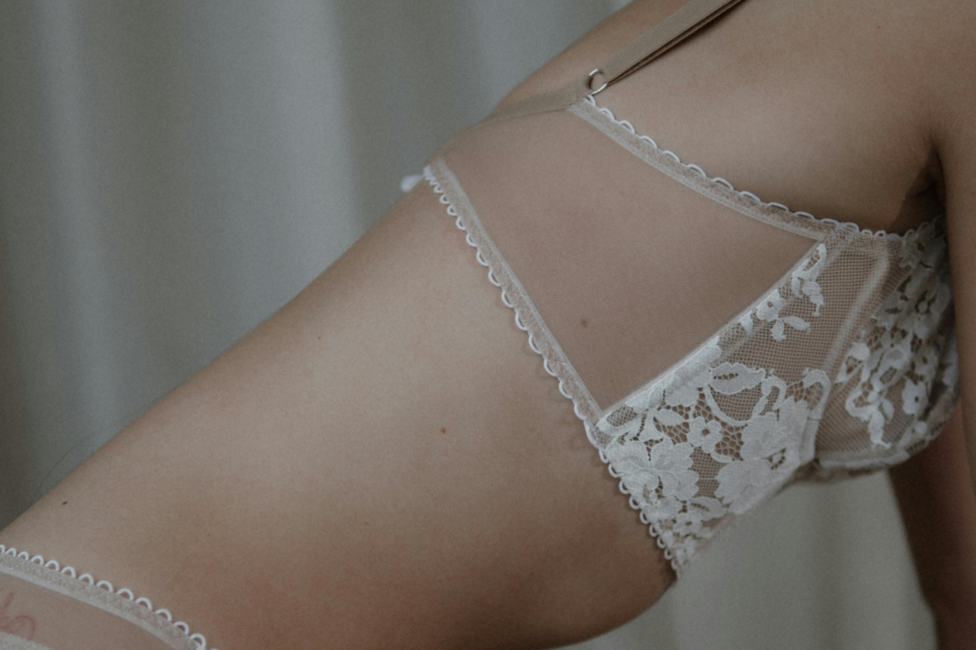A side image of a woman features diagonally against a beige background. She wears a nude and white lace bra with the lace of her underwear just showing at the bottom of the image.