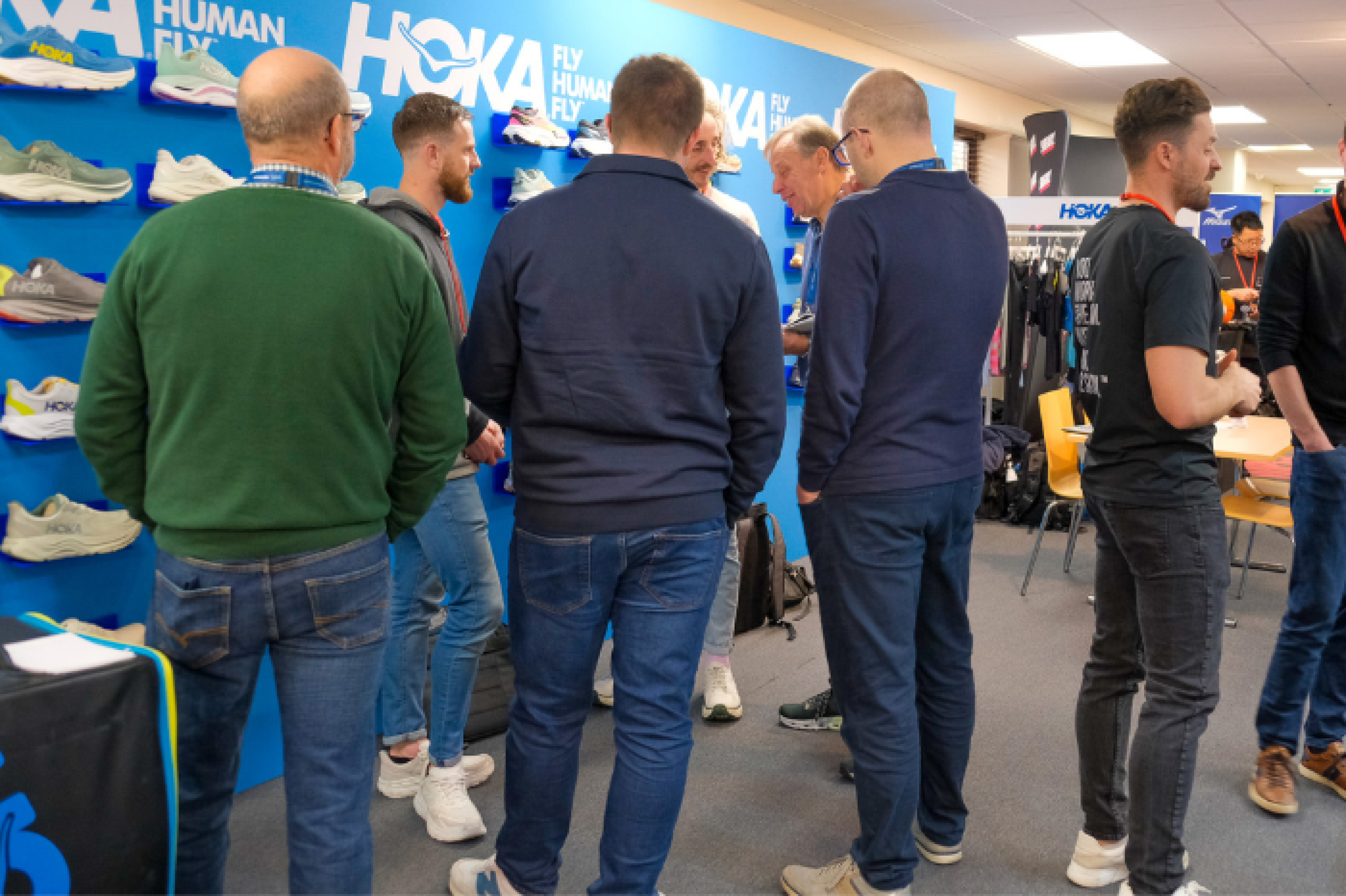 Image of retails gathering for the INDX Sports & Leisure Show at Cranmore Park. Men stand, chat, and make deals in front of a bright blue HOKA branded footwear stand at the trade show, some are wearing landyards. There is further sportswear apparel to the rear of the showroom.