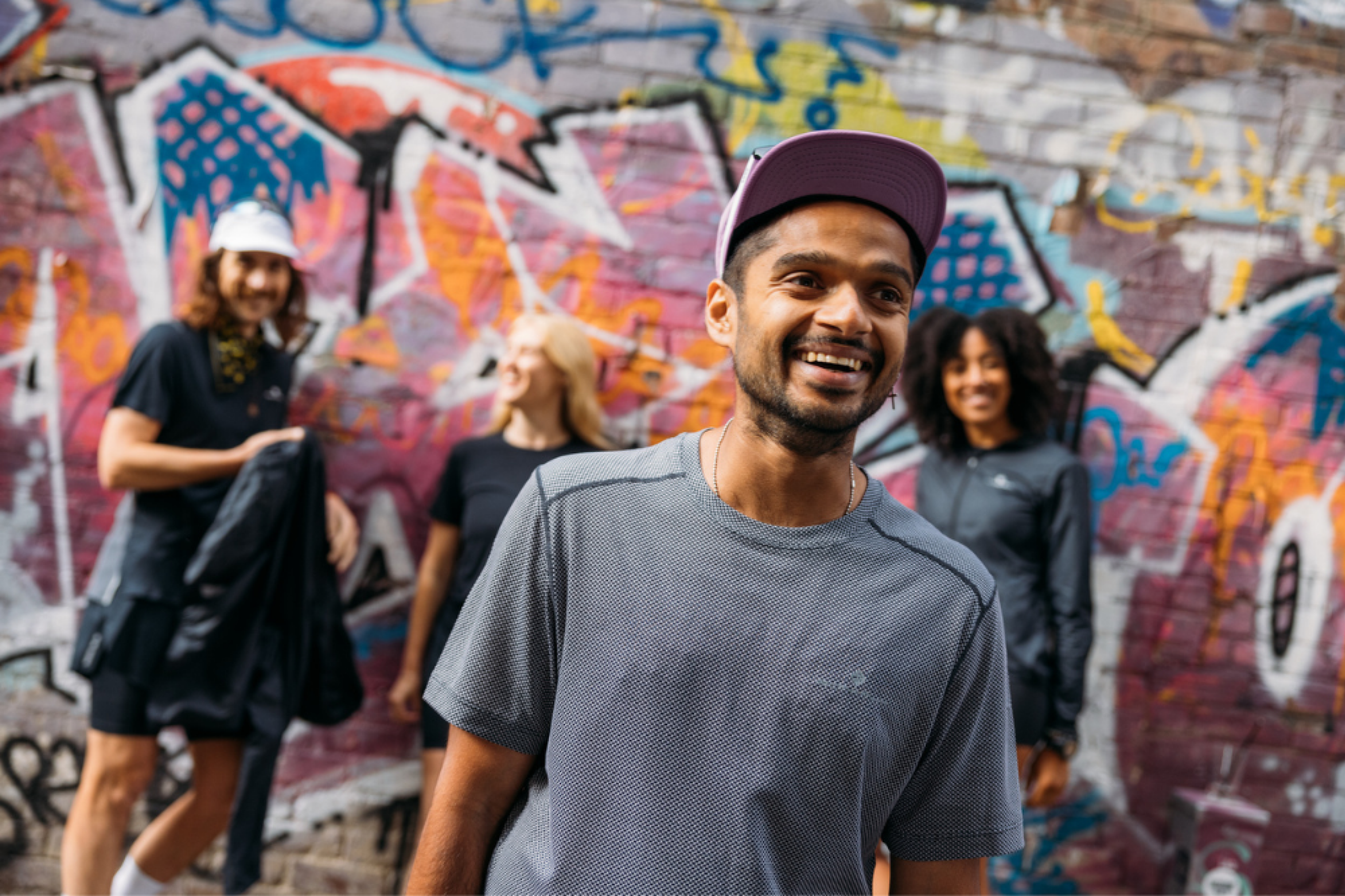 Four people stand against a colouful graffiti wall, with a man smiling forefront and central. He wears a cap and grey sports running top.