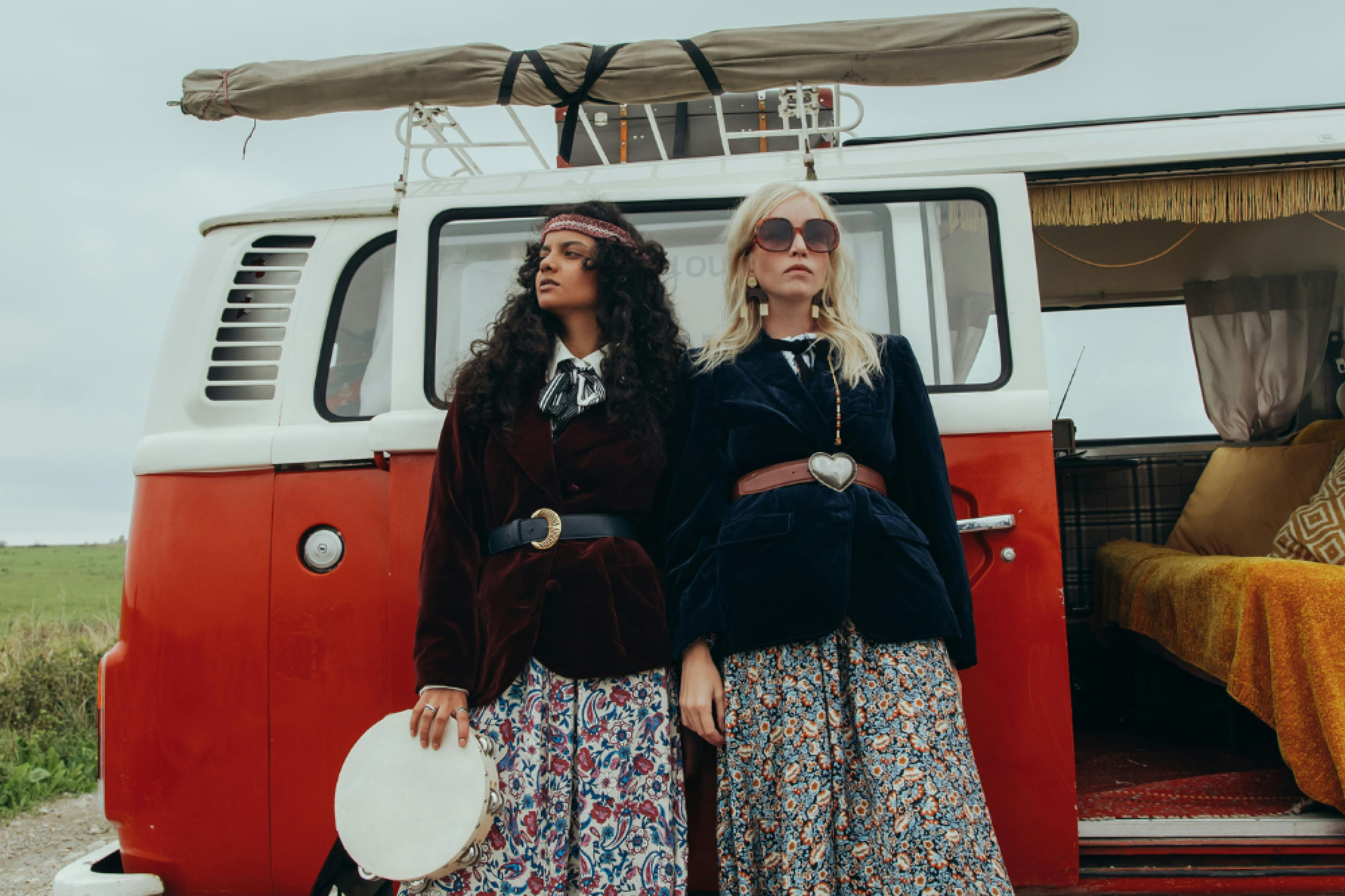 Two women feature central in floaty floral skirts, boho jackets and cinched waist belts. The woman to the left wears a headband and holds a tambourine, the woman to the right wears sunglasses. Both women stand in front of a red VW campervan with yellow furnishing seen to the right of the image.