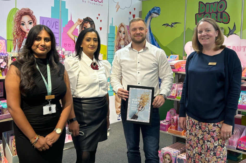 Three women and a man (centre) are standing, posed and smiling for a photo at the INDX Toy trade show. There are bold colourful brand images behind the people as well as toys and gifts for buyers to explore. The man in the centre is holding a framed Best Dressed Stand award.