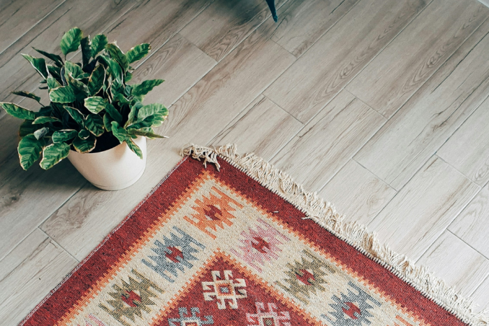 Image advertising INDX National Flooring Show. A triangle of rug features bottom central with aztec patterns in colours of orange, red, beige, blue and green. A plant to upper left of image sit on the floor in a white plantpot and laminate flooring can be seen to the background.